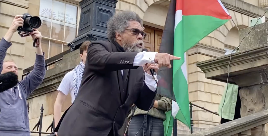 Cornel West speaking at event with Palestinian flag in background.